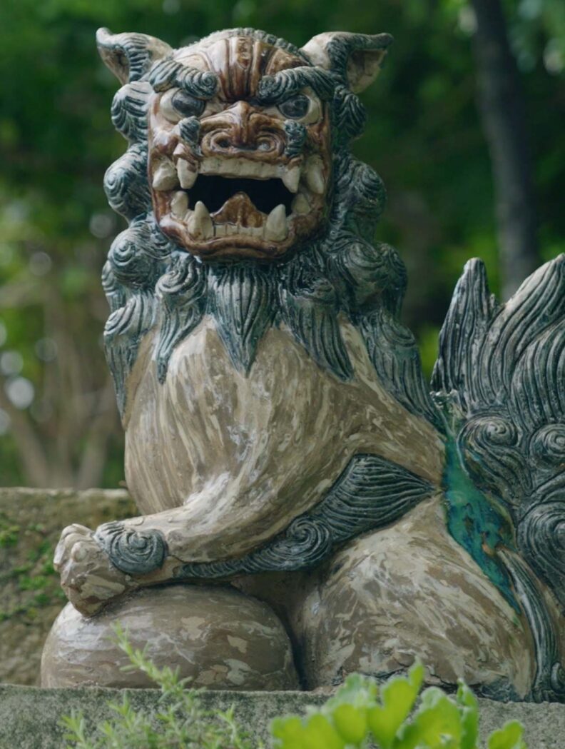 An intricately carved stone statue of a mythical lion-like creature with an open mouth showing sharp teeth, sitting on a ledge surrounded by greenery.